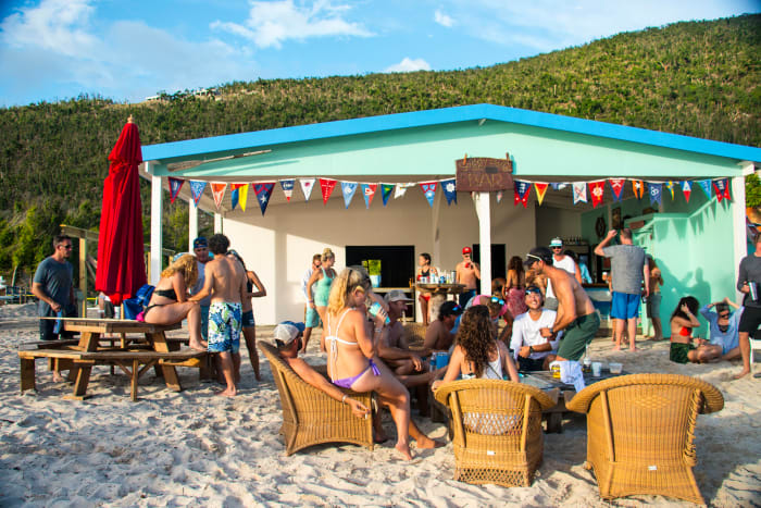 The party starts early, around noon, as visitors order drinks, swim, and relax at the tables outside The Soggy Dollar beach bar.