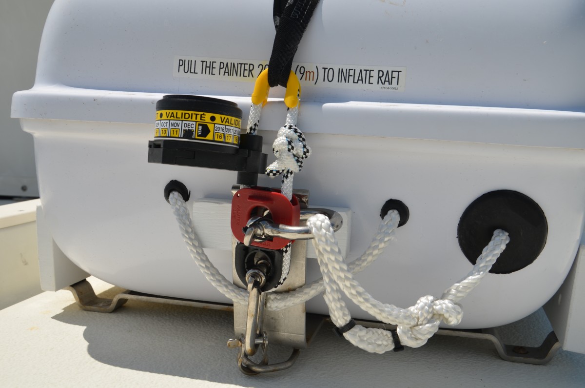A Hydrostatic Release partly consists of a strong line in an oblong circle. In this picture, it is the white line with black tracer. Instead of the life raft tie-down strap being connected directly to the cradle, it is connected instead to the loop of line at the yellow thimble at the top of the HR. The bottom of the HR device is attached to the cradle. Note the painter, which comes out of the raft at the large black rubber grommet at the right lower side; It is connected to a red “weak link” on the HR device. This particular raft has a separate white rope handle, for lifting the raft.